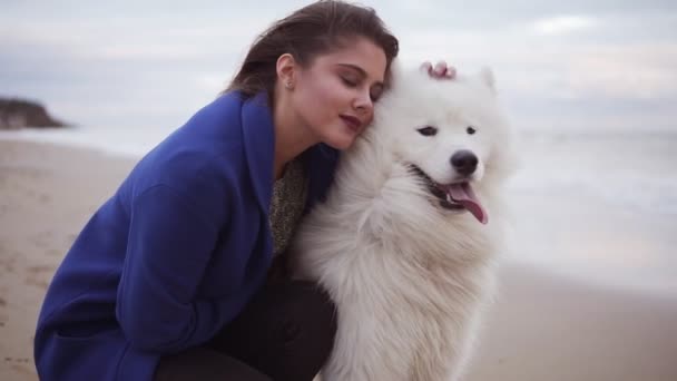 Mujer joven sentada en la arena y abrazando a su perro de la raza Samoyedo junto al mar. Blanca mascota esponjosa en la playa divirtiéndose. Hermoso cielo en el fondo. Disparo en cámara lenta — Vídeo de stock