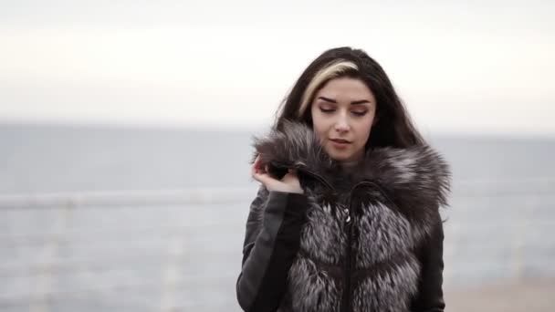 毛皮のコートを着て冬の海や海で歩く若いブルネットの女性の肖像画。彼女は近づくとカメラを探して来ています。スローモーション撮影 — ストック動画