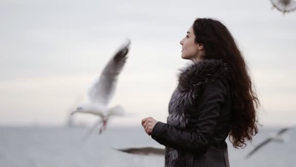 Πορτρέτο της μια ελκυστική μελαχρινή νεαρή γυναίκα στο γούνινο παλτό σίτιση οι γλάροι από τον ωκεανό το χειμώνα. Slowmotion βολή — Αρχείο Βίντεο