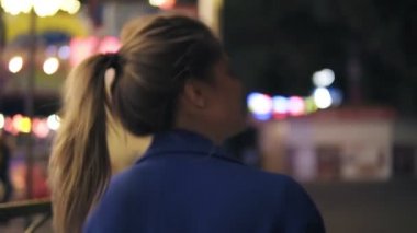 Geceleri eğlence parkında yürüyüş, etrafında dönen ve kameraya gülümseyen at kuyruğu ile arkadan görünüşü genç çekici kadın. Güzel parlak ışık arka plan yok. Slowmotion atış