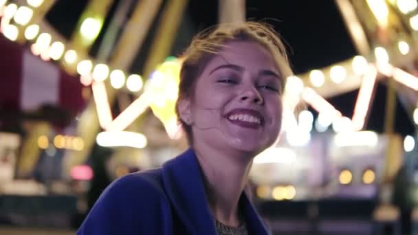 ポニーテール笑顔と背景に carusel と遊園地でぶら下がってカメラ目線を持つ美しい幸せな少女のクローズ アップ ビュー。スローモーション撮影 — ストック動画
