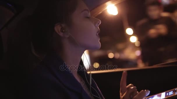 Jovem atraente está andando de táxi sentado no banco de trás e ouvindo a música em fones de ouvido, movendo as mãos, dançando, cantando sua música favorita — Vídeo de Stock