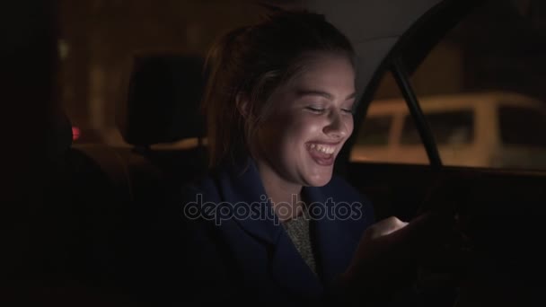 Happy smiling girl riding in a taxi at night sitting on the backseat and texting using her smartphone. Happy woman in taxi. Night live. Slowmotion shot — Stock Video