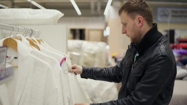 Vista de cerca de un hombre joven en una chaqueta de cuero elige un albornoz en la tienda de ropa de cama y accesorios — Vídeo de stock