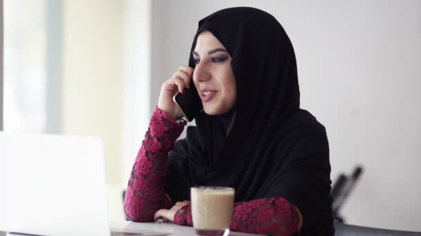 Young tersenyum bahagia muslim wanita di jilbab duduk di kafe dengan secangkir kopi dan berbicara di telepon genggamnya. Laptop modern di atas meja. Belajar atau bekerja. Perlahan-lahan — Stok Video