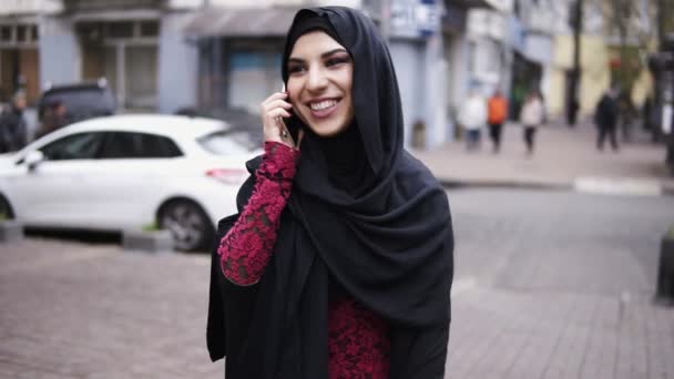 젊은 미소 매력적인 여자 그녀의 휴대 전화 통화 시에 거리에 걷는 hijab를 입고. Slowmotion 샷 — 비디오