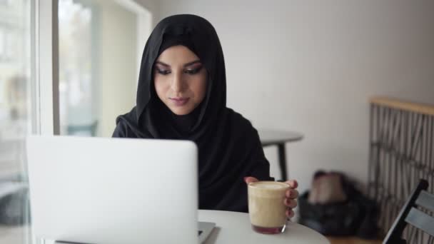 Giovane donna musulmana che lavora sul computer portatile moderno in caffè e bere cappuccino. Donna attraente in hijab alla ricerca di qualcosa in internet. Studiare o lavorare. Colpo di rallentamento — Video Stock