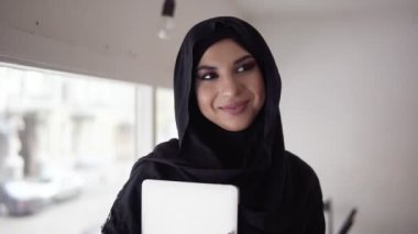 Gülümseyen ve ofis veya kafe ayakta ve onu laptop tutan kamera seyir hijab çekici kadın. Çalışma veya eğitim gülümseyen genç Müslüman kadın portresi. Slowmotion atış
