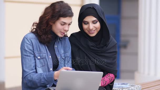 Молодая мусульманка в чёрном хиджабе разговаривает со своей кавказской подругой. Две привлекательные девушки сидят снаружи и пользуются ноутбуком. Межкультурная дружба. Слоумоушн — стоковое видео