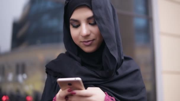 Vista de perto da jovem mulher atraente vestindo hijab em pé na rua, digitando uma mensagem em seu telefone celular. Tiro em câmara lenta — Vídeo de Stock