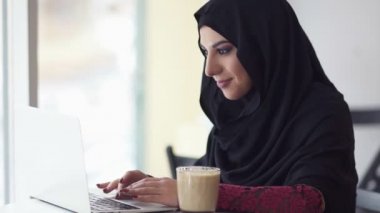 İnternet bir şey için arama hijab çekici kadın. Kafede modern dizüstü bilgisayar üzerinde çalışan ve kapuçino içmek gülümseyen genç Müslüman kadın portresi. Eğitim ya da çalışma. Slowmotion atış