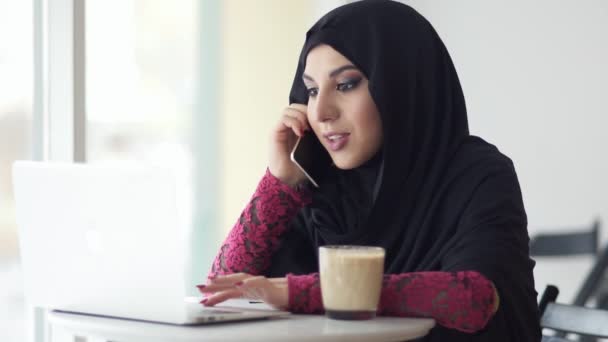 Giovane donna musulmana in hijab seduta in un caffè con una tazza di caffè e parlando sul suo cellulare mentre utilizza il suo computer portatile. Sta cercando qualcosa in internet. Studiare o lavorare. Colpo di rallentamento — Video Stock