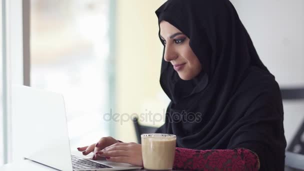 Atraktivní žena v hidžábu hledajícím v Internetu. Portrét usměvavé mladé muslimské ženy pracují na moderní notebook v kavárně a pít cappuccino. Studovat nebo pracovat. Slowmotion shot — Stock video