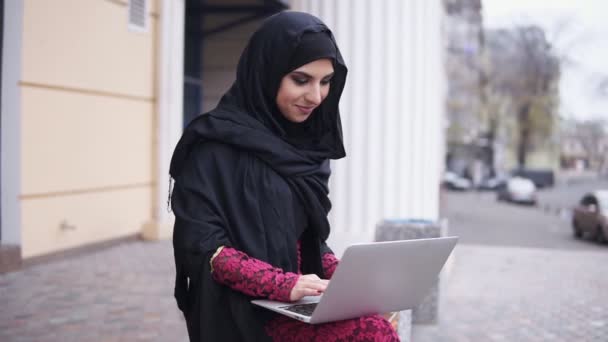 Vista de perto da mulher sorridente feliz em hijab preto sentado no banco de madeira e usando seu laptop. Estudar ou trabalhar. Tiro em câmara lenta — Vídeo de Stock