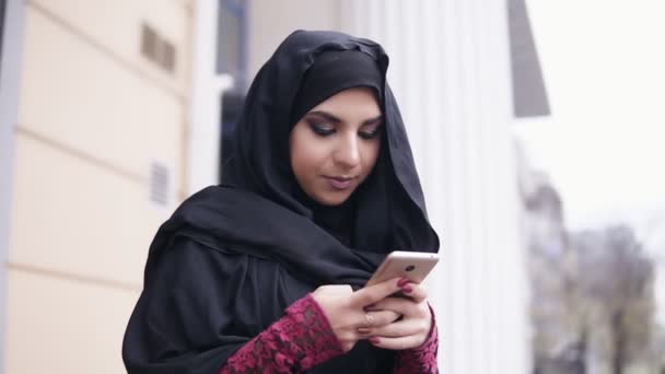 Movimento redondo da câmera: Jovem mulher atraente vestindo hijab em pé na rua, digitando uma mensagem em seu telefone celular. Tiro em câmara lenta — Vídeo de Stock