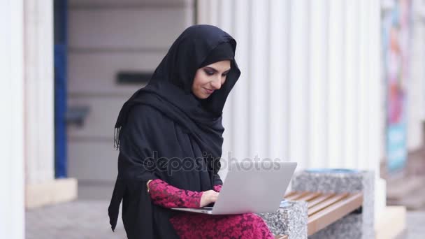 Giovane donna in hijab nero seduto in strada e alla ricerca di qualcosa in internet. Ritratto di giovane donna musulmana sorridente che lavora su un computer portatile moderno per strada. Studiare o lavorare. Rallentamento — Video Stock