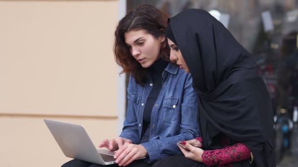 Дві жінки сидять на вулиці і використовують ноутбук. Перехресна культурна дружба. Молода мусульманка в чорному хіджабі розмовляє зі своїм жіночим кавказьким другом. Постріл уповільнення — стокове відео