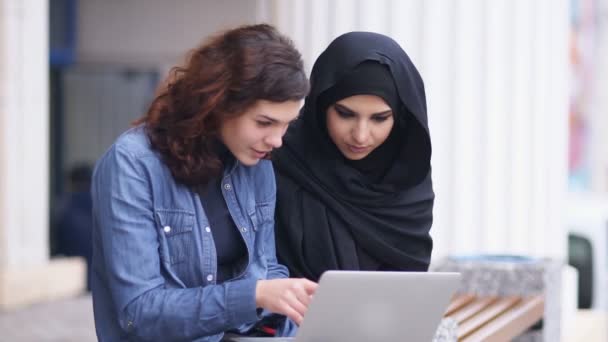 Interculturele vriendschap. Jonge moslimvrouw in zwarte hijab is in gesprek met haar blanke vriendin. Twee aantrekkelijke jonge vrouwen zitten buiten en het gebruik van de laptop. Tikje schot — Stockvideo