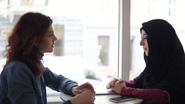 Dos amigas internacionales hablando en la cafetería: una joven musulmana en hiyab negro está hablando con su amiga caucásica mientras está sentada en la cafetería. Disparo en cámara lenta — Vídeo de stock