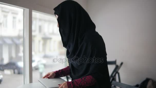 Ελκυστική γυναίκα στο χιτζάμπ με σύγχρονο laptop κάθεται στο τραπέζι και αρχίζει να λειτουργεί. Slowmotion βολή — Αρχείο Βίντεο