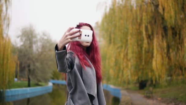 年轻迷人的女人与红色停顿做自拍在她的智能手机, 而站在一个人工池塘在一个秋季公园 — 图库视频影像