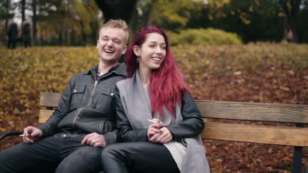 Junges attraktives Paar sitzt auf einer Bank im Herbstpark und unterhält sich. attraktive Frau mit roten Haaren raucht — Stockvideo