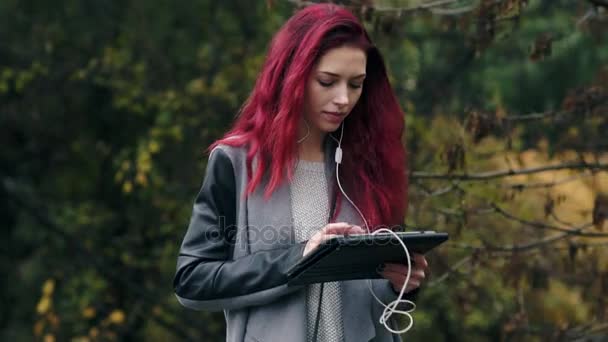 Jovem mulher bonita com cabelos vermelhos caminha no parque de outono e ouve música via tablet pc. Jovem mulher atraente com computador tablet digital nas mãos — Vídeo de Stock