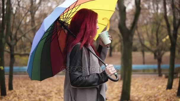 Vue de côté : jeune femme aux cheveux roux marchant dans le parc d'automne et buvant du café dans une tasse en papier tout en tenant un parapluie coloré. Fille en manteau chaud profiter du temps frais d'automne avec une tasse de boisson chaude 