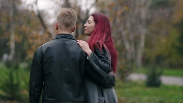 Romantic young couple walking in autumn park during the day. Back view of young blonde man in leather jacket embracing his girlfriend. Loving couple talking and spending time together — Stock Video