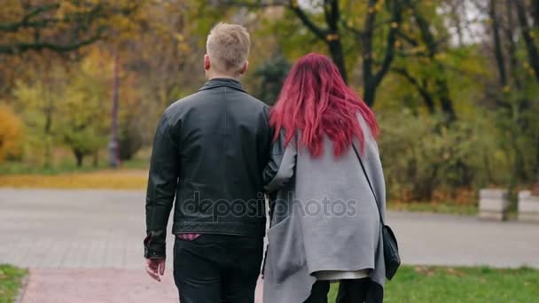 BaCl weergave van romantische jonge paar wandelen in de herfst park tijdens de dag. Achteraanzicht van jonge blonde man in leren jack en zijn vriendin praten en tijd samen doorbrengen — Stockvideo