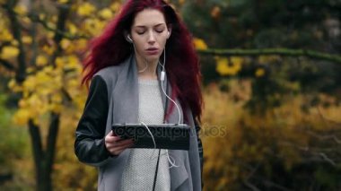 Kızıl saçlı genç işkadını sonbahar parkta yürüyor ve tablet pc yolu ile iletişim kurar. Dijital tablet bilgisayar elinde olan çekici kadın kulaklık kullanarak iletişim kurar