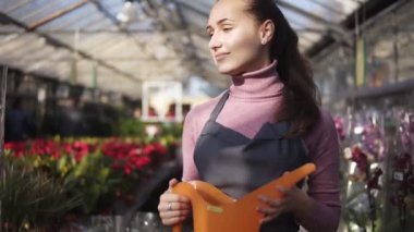 Tek tip bir sera raflara farklı bitkiler arasında yürüyüş ve turuncu sulama olabilir onun elinde tutan genç kadın bahçıvan