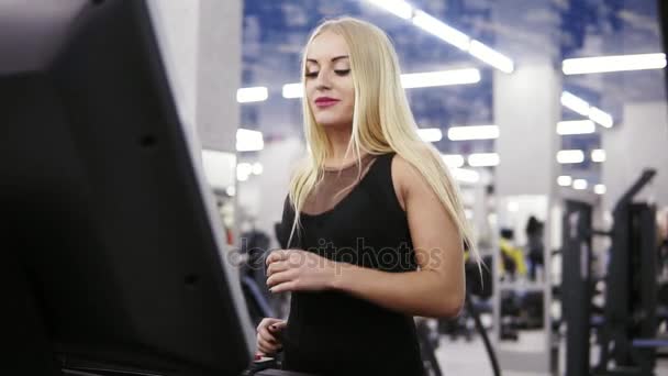 Молода приваблива біла жінка з світлим волоссям в чорному спортивному вбранні біжить на біговій доріжці в спортзалі — стокове відео