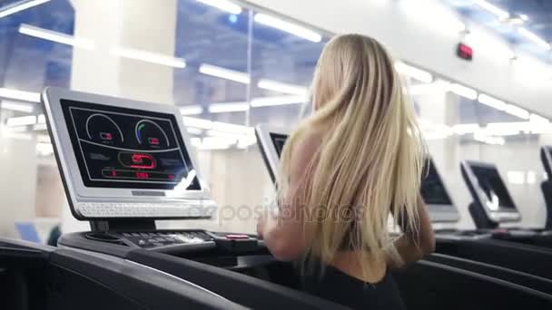 Joven atractiva mujer caucásica con cabello rubio en traje deportivo negro corriendo en treadmil en el gimnasio en slomo — Vídeos de Stock