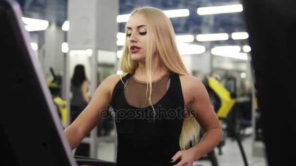 Joven atractiva rubia caucásica con piercing en traje deportivo negro respirando intensamente mientras trota en simulador de cinta de correr en slomo — Vídeos de Stock