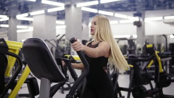 Joven mujer rubia atractiva sonriendo mientras pedalea en simulador de entrenador elíptico en traje deportivo negro en el gimnasio. Cuidado de la salud, fitness, culturismo . — Vídeos de Stock