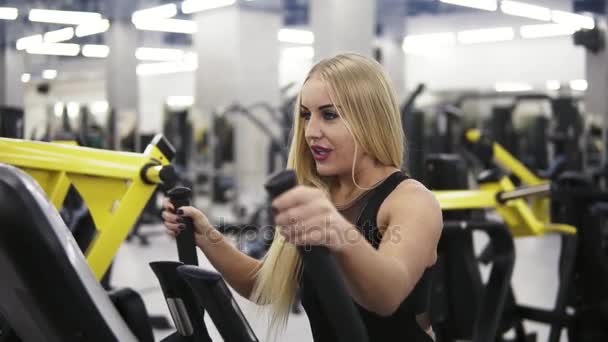 Mulher sexy nova bonita com cabelo loiro em roupa de esporte preto sorrindo enquanto trainig no simulador de treinador elíptico no ginásio. Fitness, bem-estar, cuidados com o coração . — Vídeo de Stock