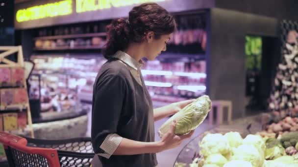 Jonge mooie brunette meisje in haar 20s plukken uit napa kool en bloemkool en brengen hen in winkelwagentje op de groenten- en fruitsector gangpad in een supermarkt — Stockvideo