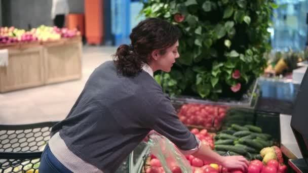 Joven chica morena hermosa en sus años 20 recogiendo tomates en una bolsa de plástico en el pasillo de frutas y verduras en una tienda de comestibles — Vídeos de Stock