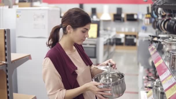 Jonge vrouwelijke klant cook pot kiezen uit allerlei keukengerei in een toestel winkel. Behandeling van carefuly staal pannen in een rij showcase. — Stockvideo