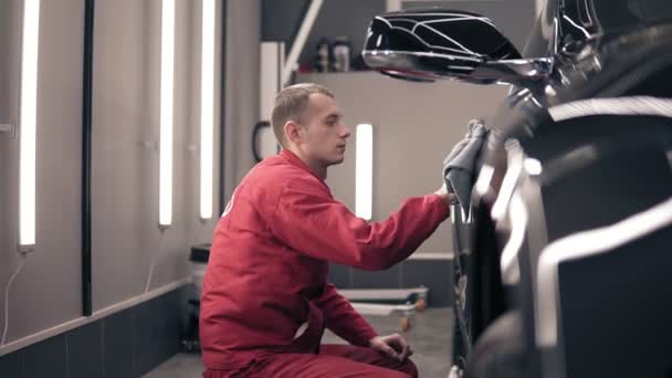 Man in rode worksuit wrijft zwarte expesive auto met de Wisser van microfiber. Polijsten op AutoCentreren (AutoCenter). — Stockvideo