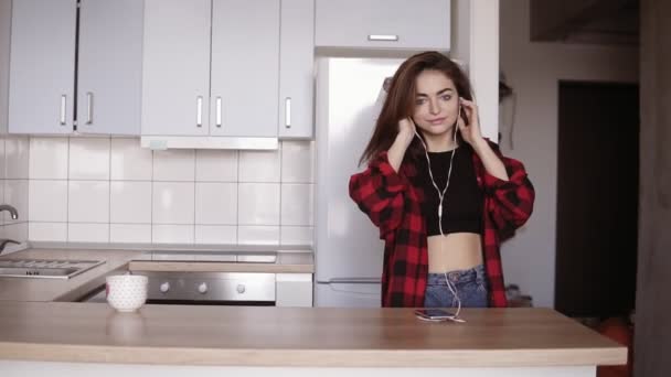 Молодая сексуальная девушка в возрасте 20 лет танцует на кухне с наушниками в ушах, берет чашку со стола . — стоковое видео