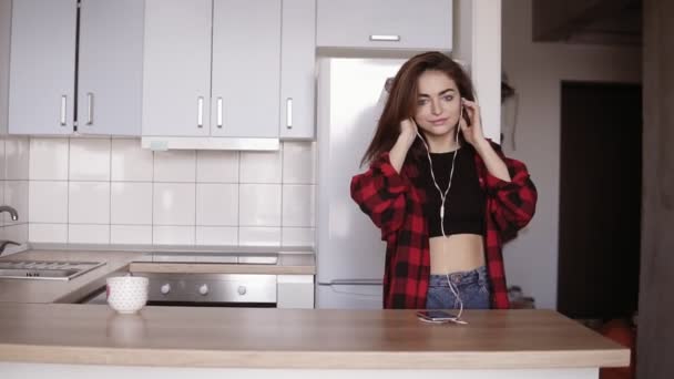 Joven chica sexy en sus años 20 bailando en la cocina con auriculares en los oídos, toma una taza de la mesa . — Vídeo de stock