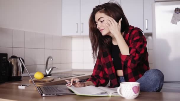 Chica atractiva en sus 20 años busca algo en Internet usando su computadora portátil y luego escribe algo en su cuaderno mientras está sentada en una acogedora cocina de su apartamento — Vídeo de stock