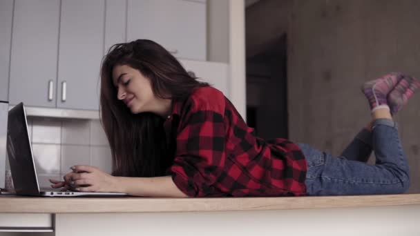 Una chica de 20 años acostada en la superficie de la mesa de la cocina y escribiendo algo en el portátil . — Vídeos de Stock