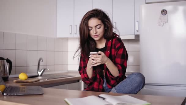 Belle fille brune texter quelqu'un tout en étant assis dans la cuisine de son appartement . — Video