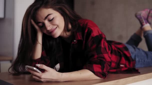 Menina morena atraente está sonhando e sorrindo sobre algo enquanto deitado na superfície da mesa da cozinha e enviando mensagens de texto para alguém. Imagens de perto . — Vídeo de Stock