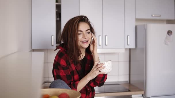 Mooi meisje begint iemand te roepen terwijl zittend in haar keuken en met een kopje thee in haar hand. — Stockvideo