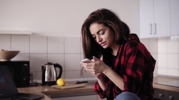 Menina morena bonita nova com cabelo bagunçado na camisa de flanela sentada em sua cozinha e enviando mensagens de texto a alguém . — Vídeo de Stock