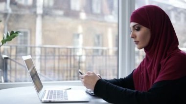 Güzel genç kadın, mor hijab kafasını kapsayan bir ışık tanınmaz kapalı alan içinde belgili tanımlık laptop yanında oturan onun smartphone, kayar